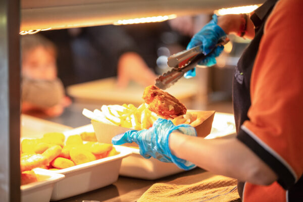 A person wearing blue gloves serving fish and chips using tongs under a warm light.