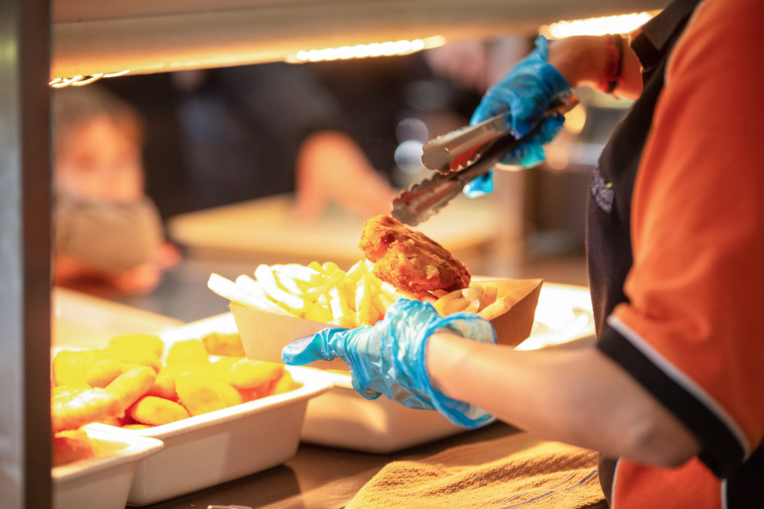 A person wearing blue gloves serving fish and chips using tongs under a warm light.