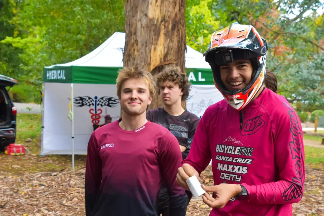 Three young men pose outdoors in a wooded area. One, in a red shirt and helmet, smiles broadly. A medical tent and trees are visible in the background.