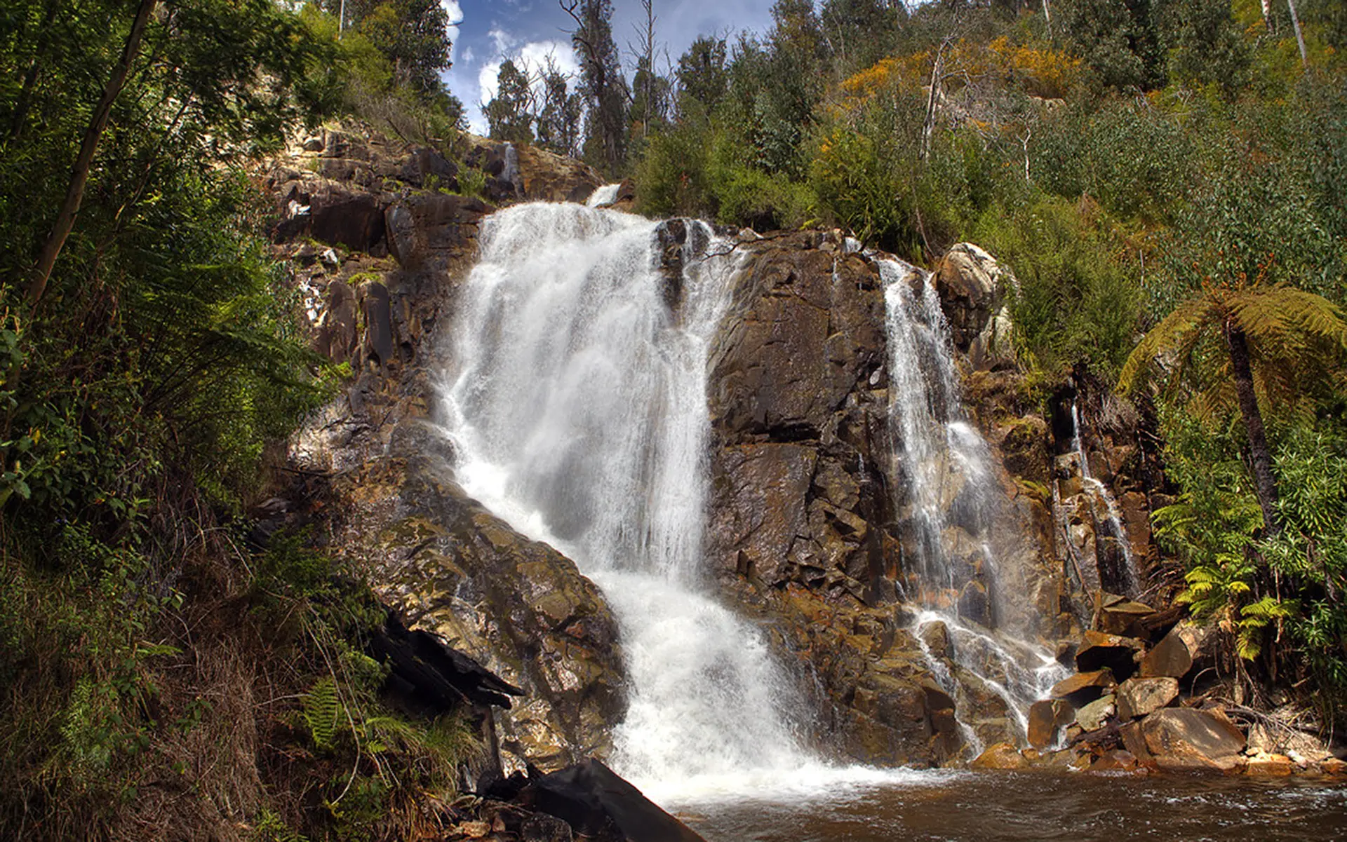 A tranquil waterfall cascades over rugged rocks, surrounded by lush green foliage and trees under a partly cloudy sky, conveying a serene, natural beauty.